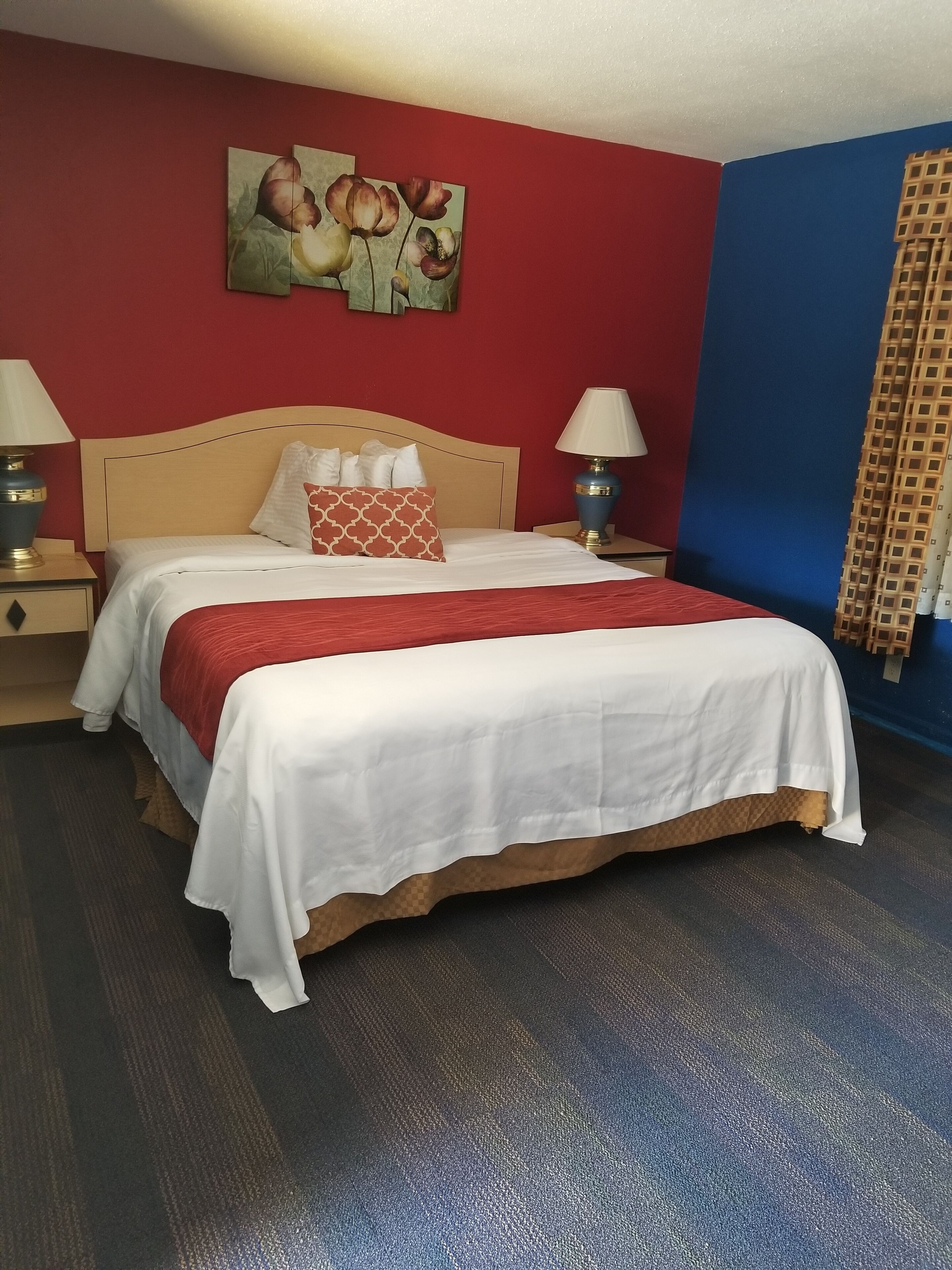 A neatly made bed with white and red bedding, two bedside lamps, and a red and blue wall with a floral painting above the bed.