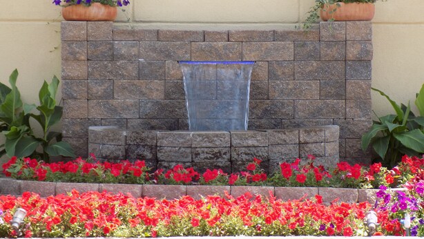 Un mur de pierre agrémenté d’un élément décoratif avec de l’eau et de parterres de fleurs.