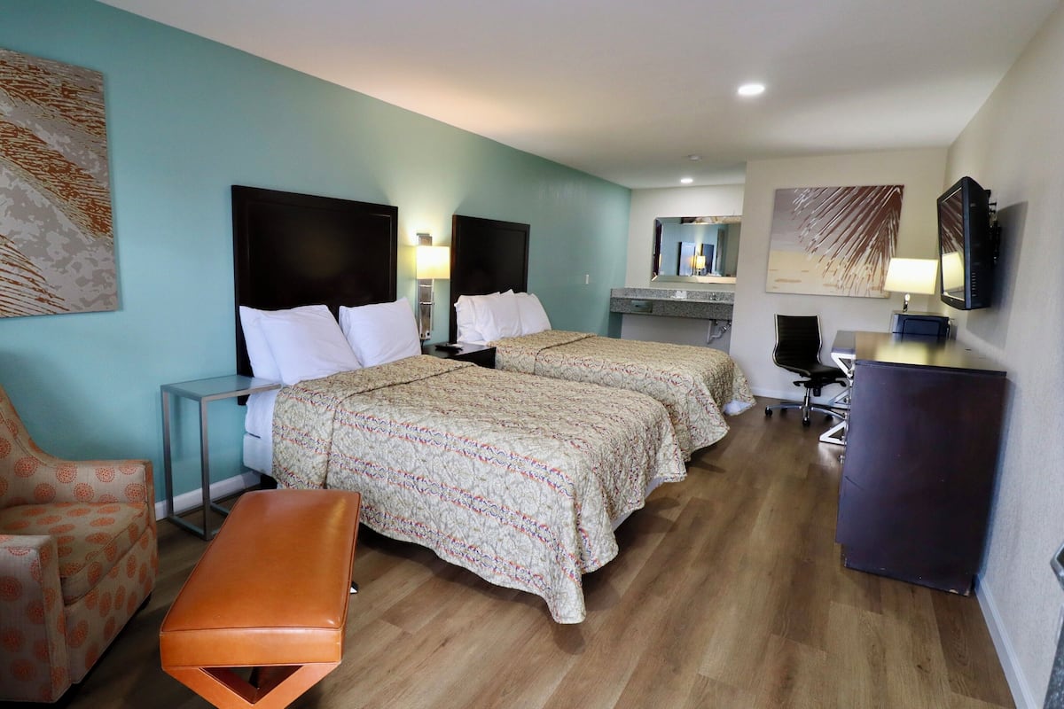 Quarto de hotel com duas camas, uma mesa, uma cadeira, um banquinho e obras de arte nas paredes.