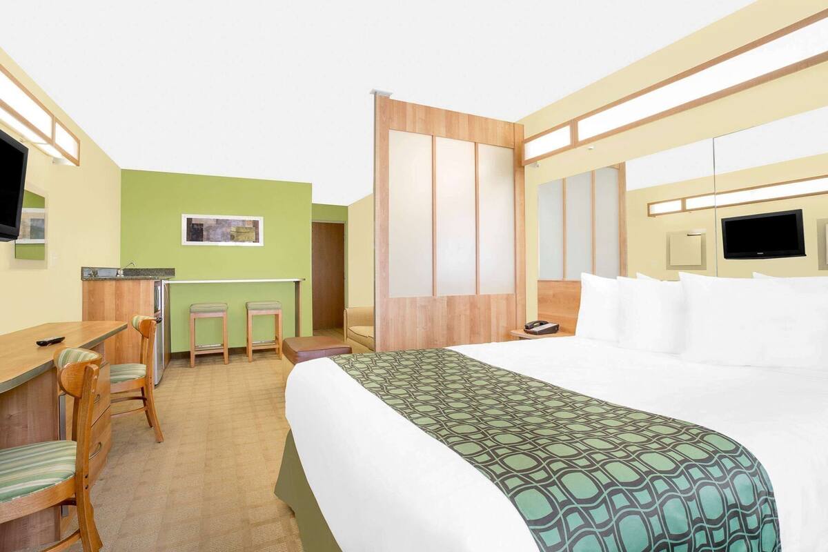 A hotel room with a bed, a desk with chairs, a small kitchenette, and a TV.