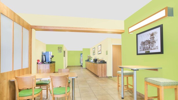 A dining area with wooden tables and chairs, a kitchen in the background, and a framed picture on the wall. - Microtel Inn & Suites by Wyndham Tuscumbia/Muscle Shoals (Tuscumbia)