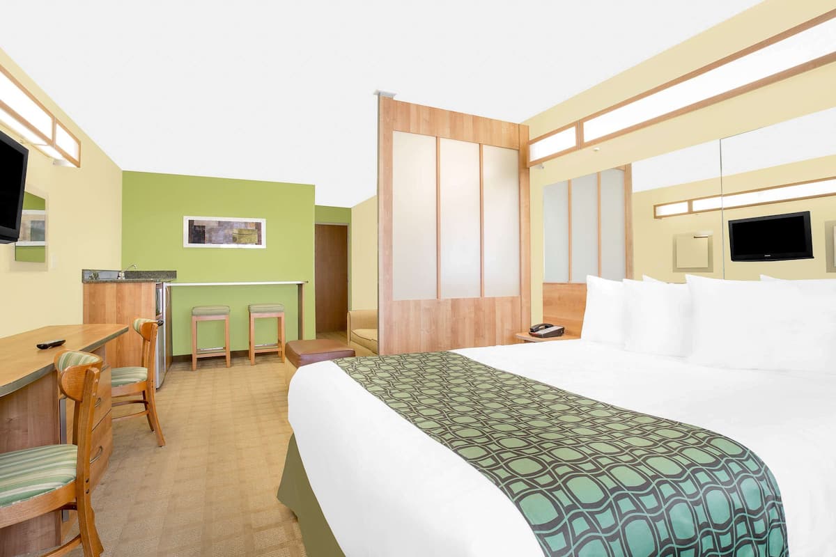 A hotel room with a bed, a desk with chairs, a small kitchenette, and a TV.