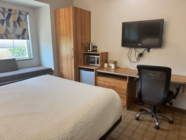 A hotel room with a bed, a desk with a chair, a TV, a microwave, and a small refrigerator.