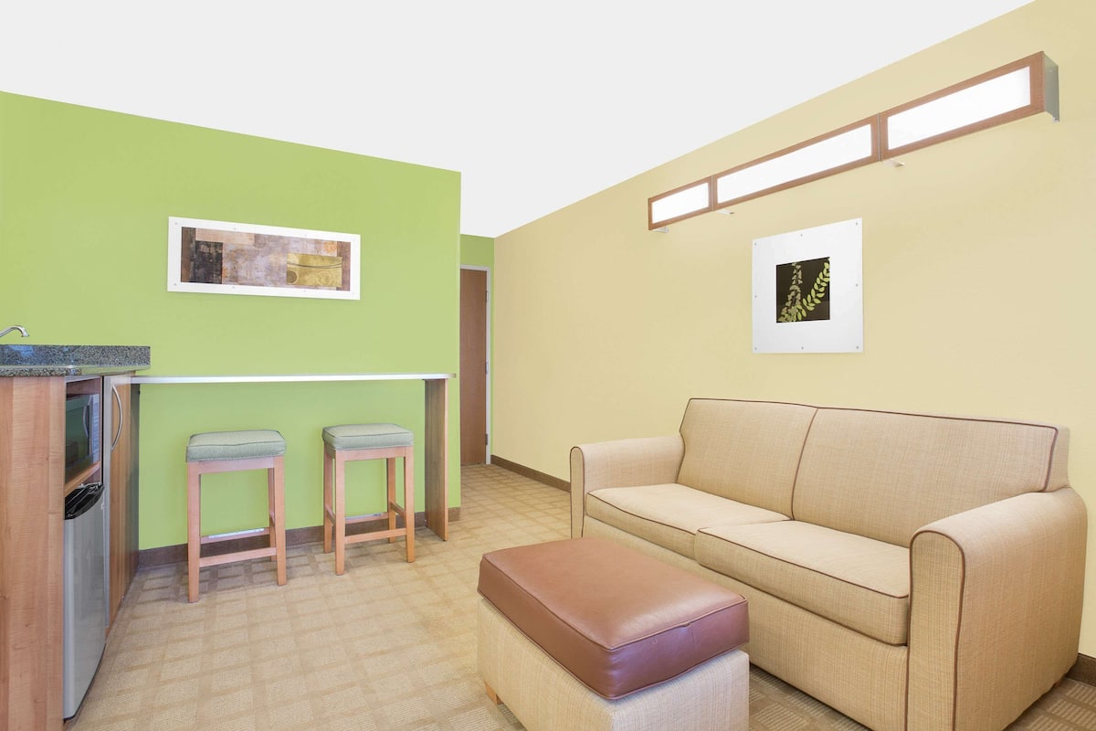 A living room with a sofa, two stools, a small table, and a kitchenette area.