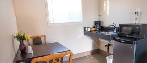 A compact kitchen with a microwave, sink, and a small refrigerator. There is a dining table with chairs and a vase with flowers.