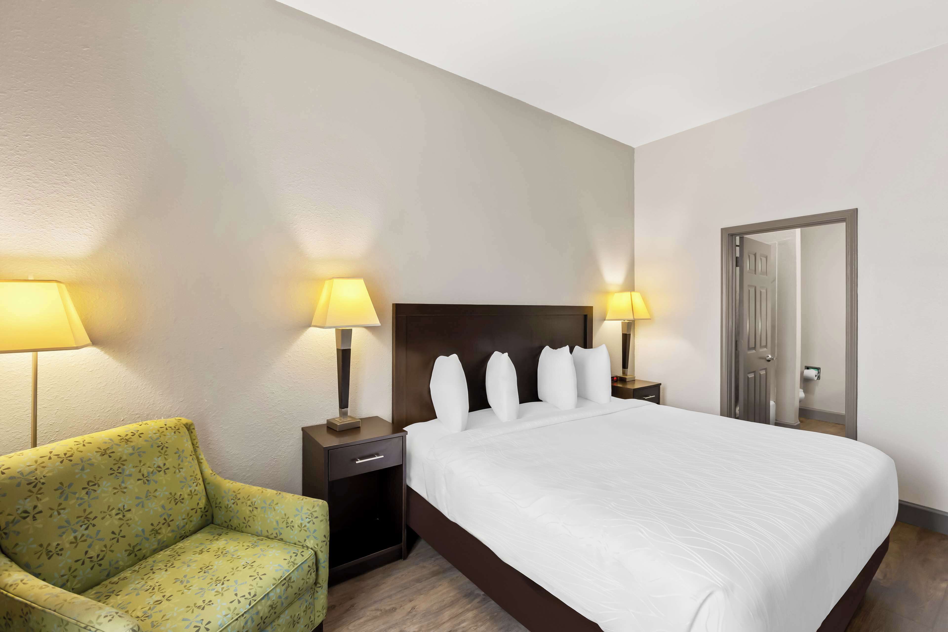 A hotel room with a large bed, two bedside lamps, a nightstand, a floral-patterned armchair, and a door.