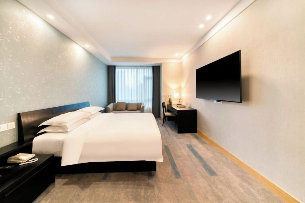 A hotel room with a large bed, a flat-screen TV mounted on the wall, a desk with a chair, and a window with curtains.