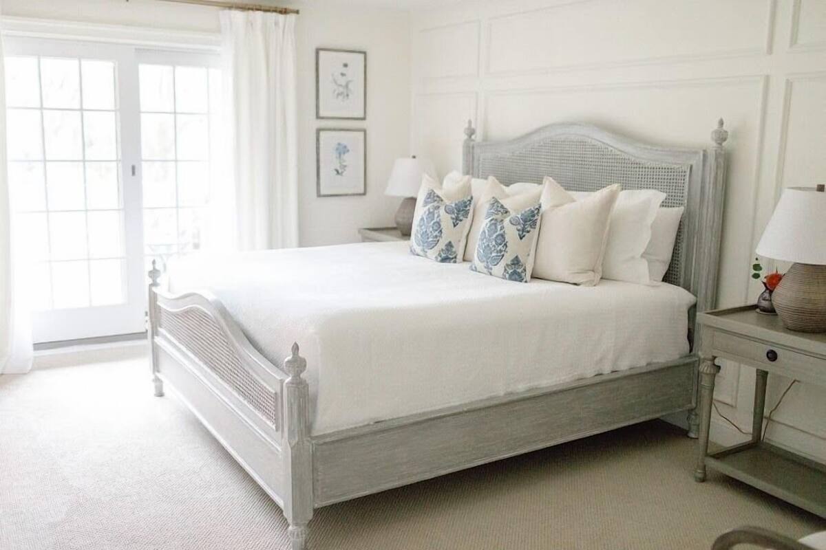 A white four-poster bed with a grey headboard, white bedding, and blue floral pillows. There are two bedside tables with lamps and a framed picture on the wall.