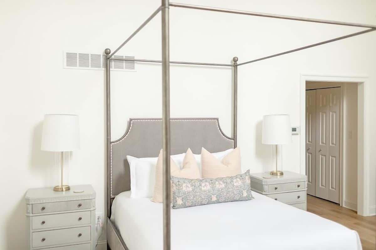A four-poster bed with a grey headboard, two bedside tables with lamps, and a patterned throw pillow.