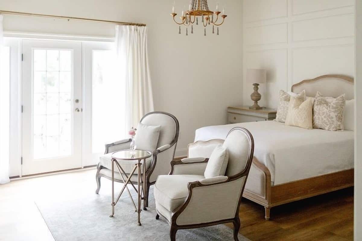 A bedroom with a bed, two armchairs, a small round table, a chandelier, and a nightstand with a lamp.