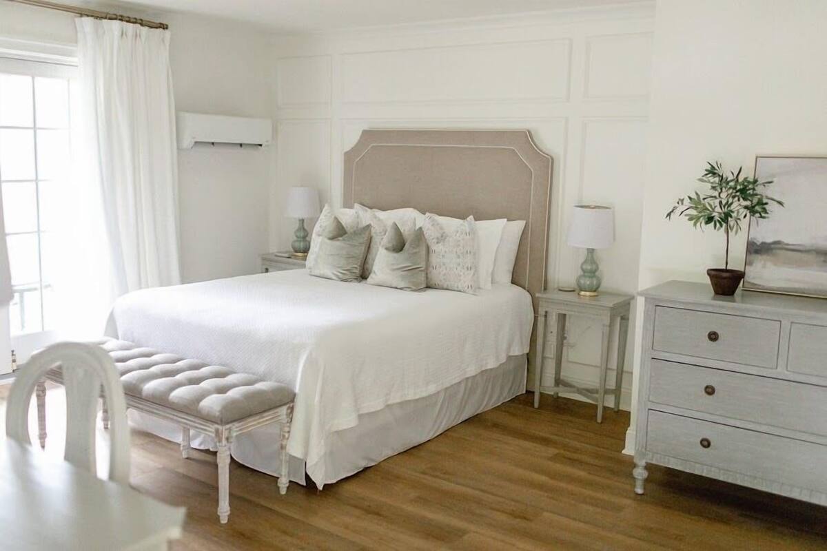 A bedroom with a large bed, a bedside table with a lamp, a dresser, and a chair.