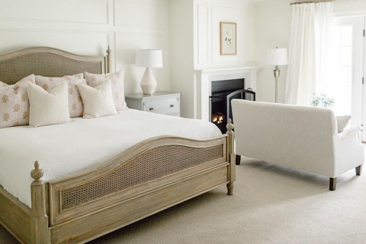 A bedroom with a large bed, a white armchair, a nightstand, a bedside table, a fireplace, and a framed picture on the wall.