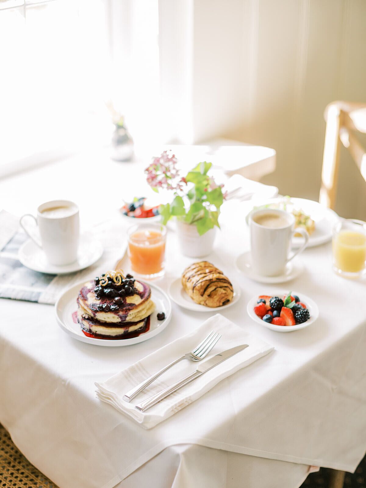 A breakfast table with pancakes, a croissant, fresh berries, a glass of orange juice, and a cup of coffee.