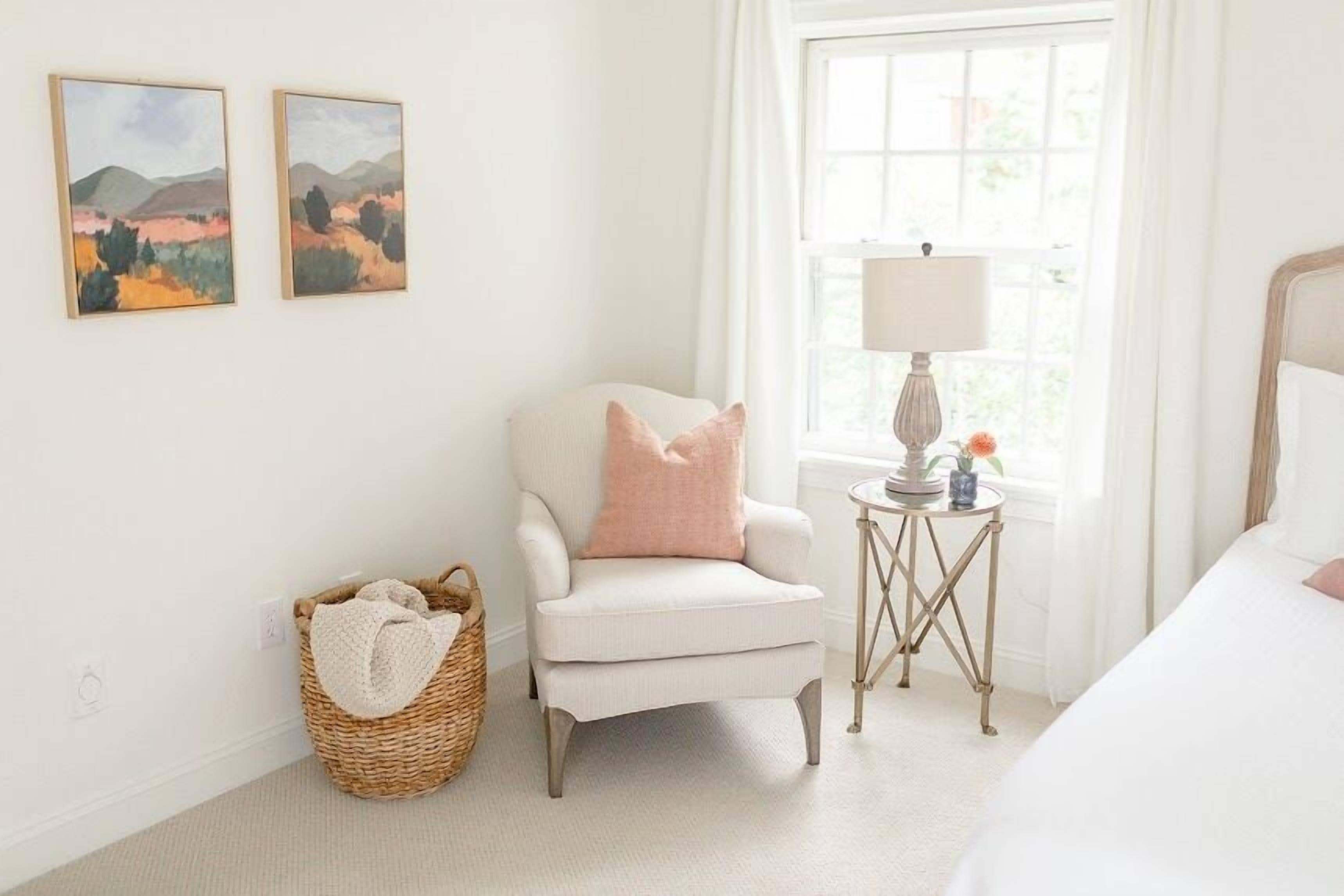 A bedroom with a white armchair, a bedside table with a lamp, and two framed landscape paintings on the wall.