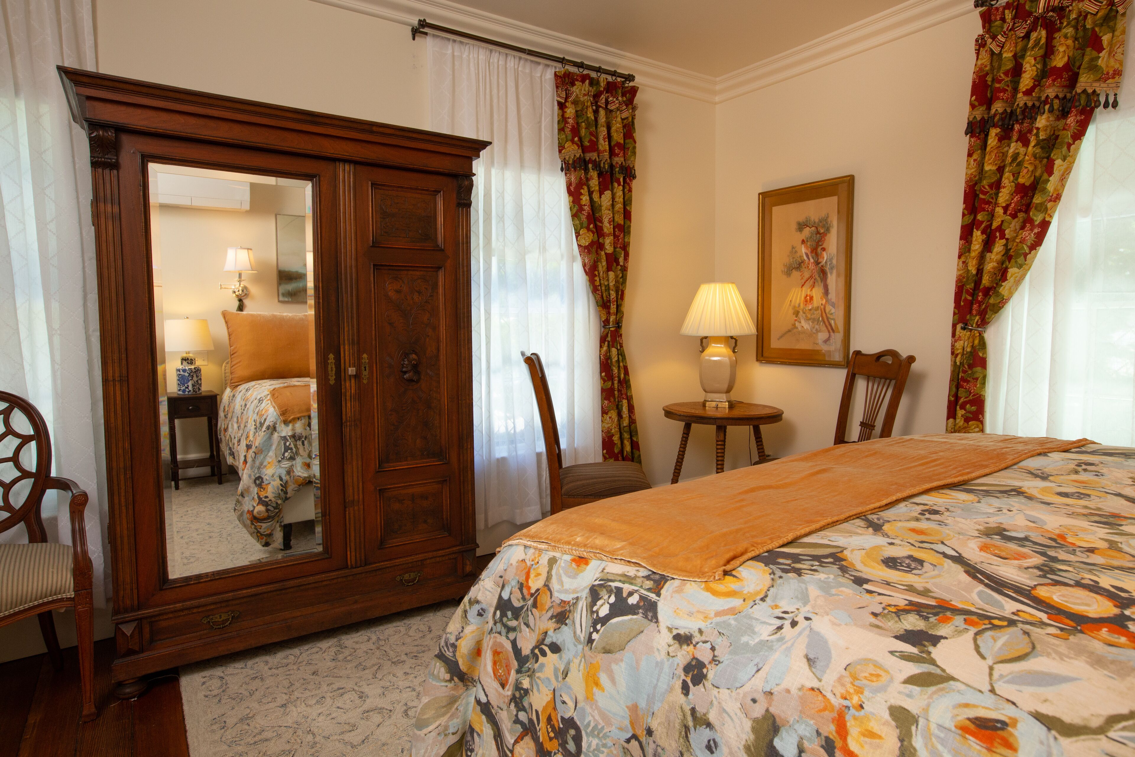 Una camera da letto con un grande specchio, un letto con copripiumino floreale, un armadio in legno, una sedia, un tavolino con una lampada e un quadro appeso al muro.