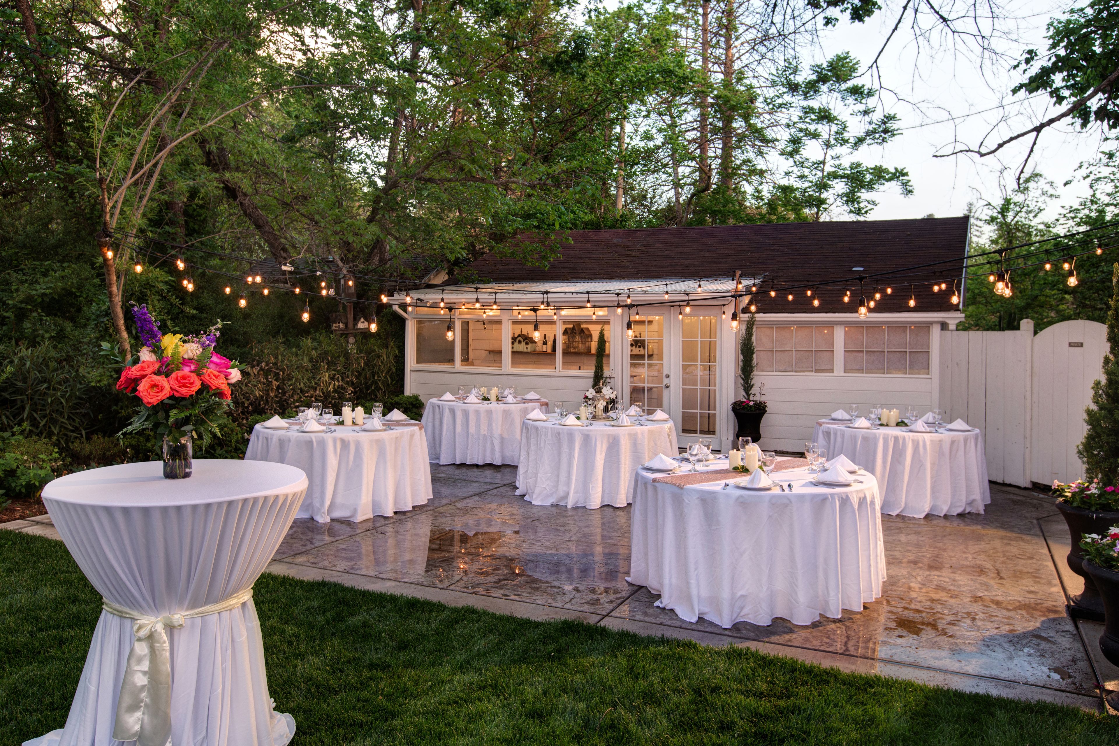 An outdoor patio with round tables set for dining, surrounded by greenery and string lights.