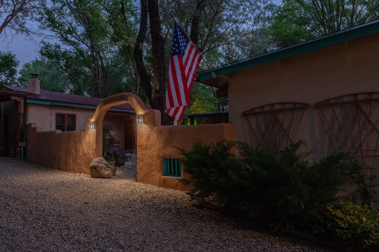 A rustic house with a distinctive arched entrance, an American flag, and a gravel driveway.