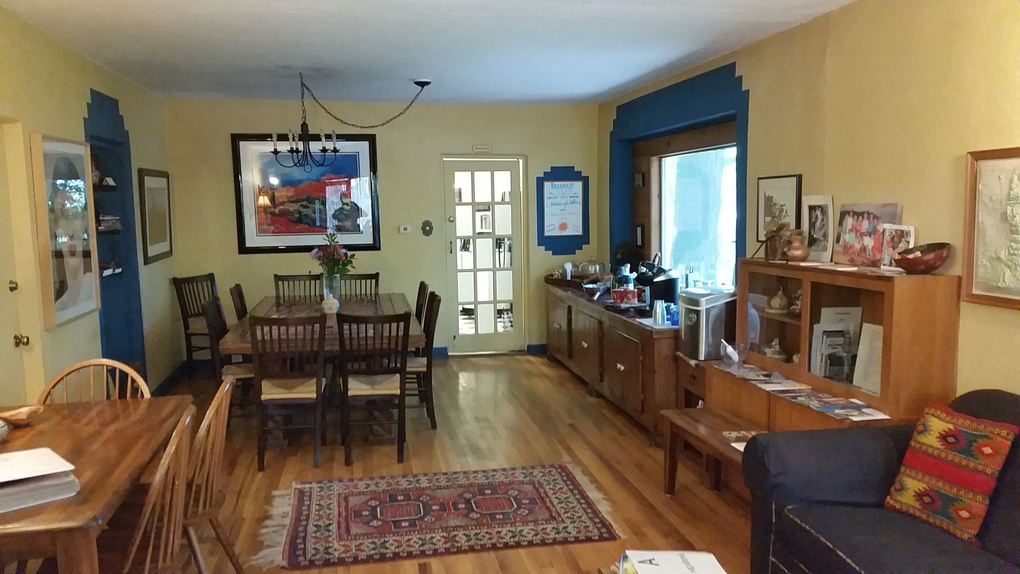 A dining area with a wooden table and chairs, a chandelier, and a rug. There is a kitchen area with a stove and cabinets.