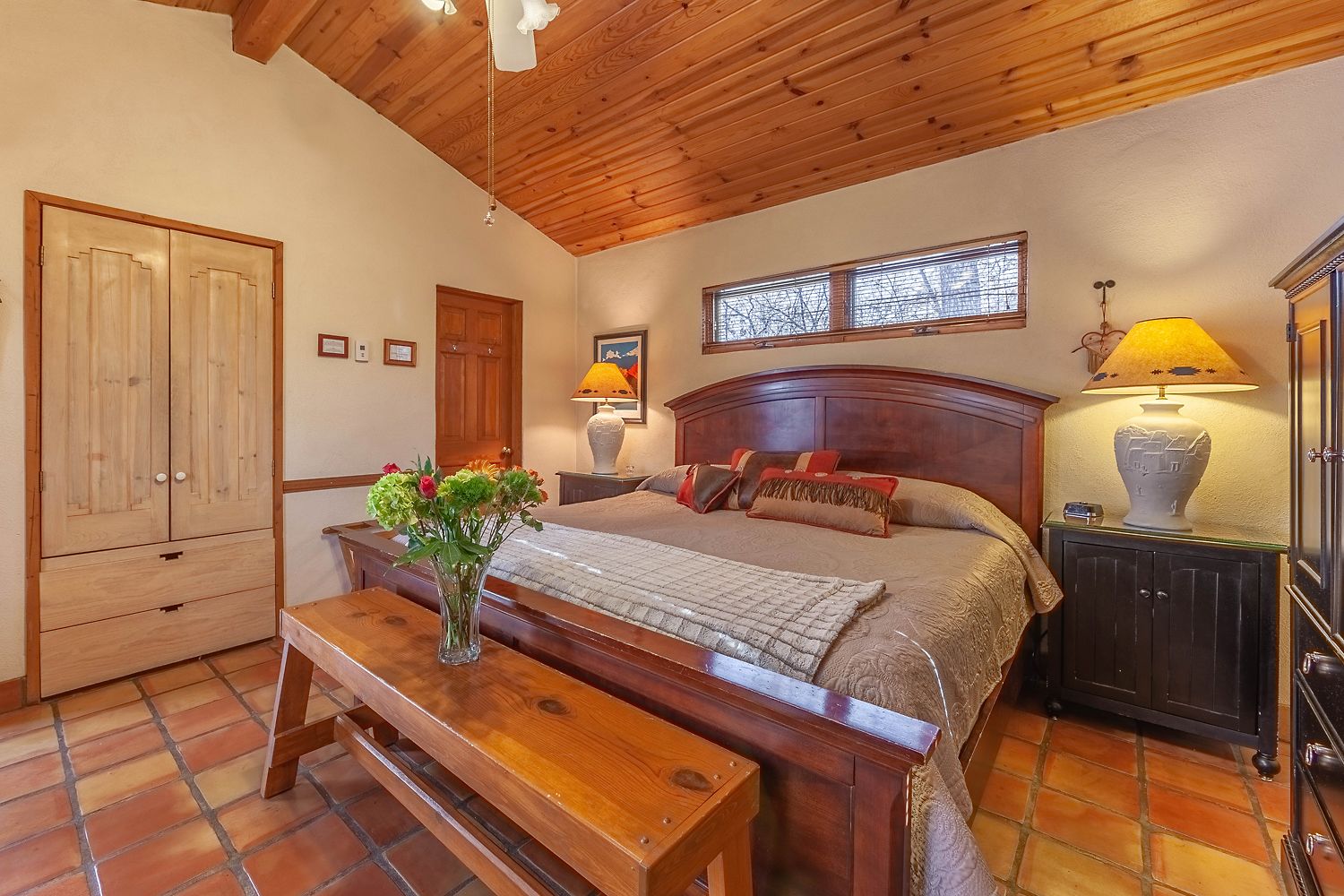 A bedroom with a wooden ceiling, a bed with multiple pillows, a nightstand with a lamp, a wooden wardrobe, and a vase with flowers on a table.