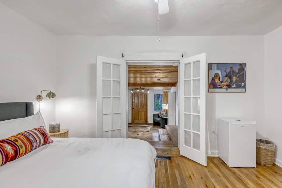 A bedroom with a bed, a nightstand, a white refrigerator, and an open door leading to a hallway.