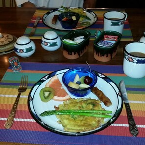 Mesa com um jogo americano colorido, um prato com comida que inclui uma fatia de kiwi, aspargos grelhados e uma pequena tigela de fruta. Também há duas xícaras com um desenho, uma faca e um garfo sobre a mesa.