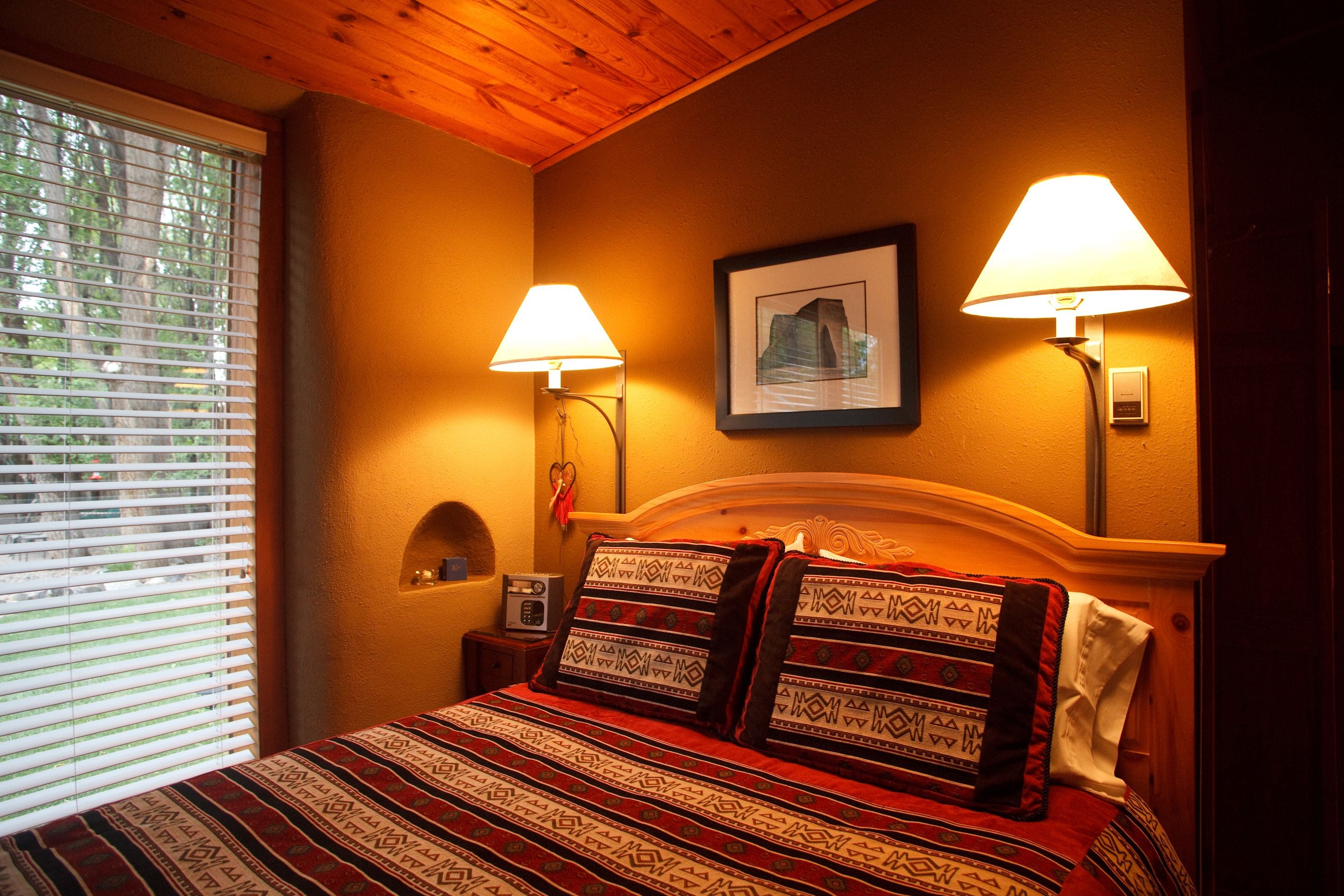 A bedroom with a wooden headboard, two bedside lamps, a framed picture on the wall, and a window with blinds.