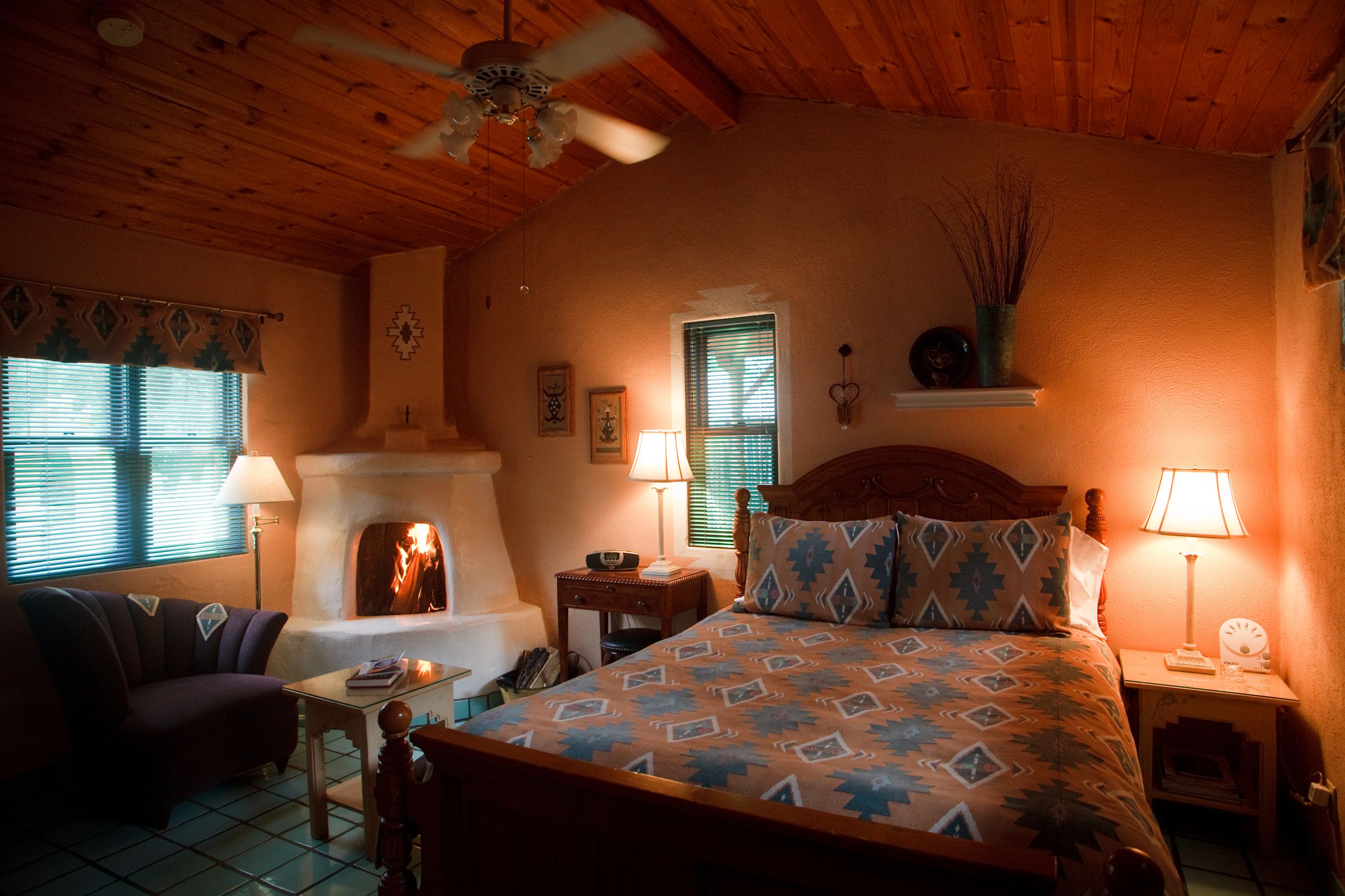 A cozy room with a fireplace, a bed with patterned bedding, a wooden ceiling, and a nightstand with a lamp.