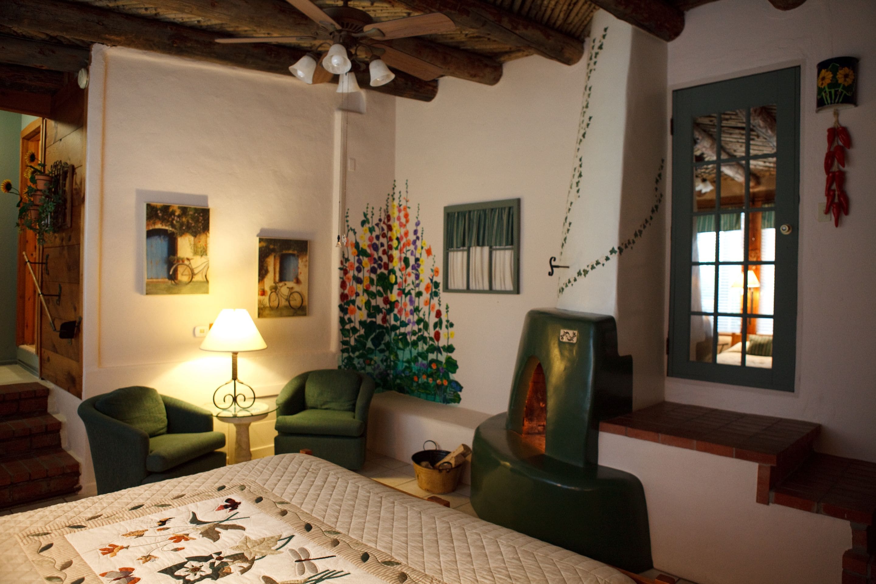 Une pièce chaleureuse avec un lit, deux fauteuils verts, une lampe, un mur orné d’une fresque colorée et un plafond en bois.