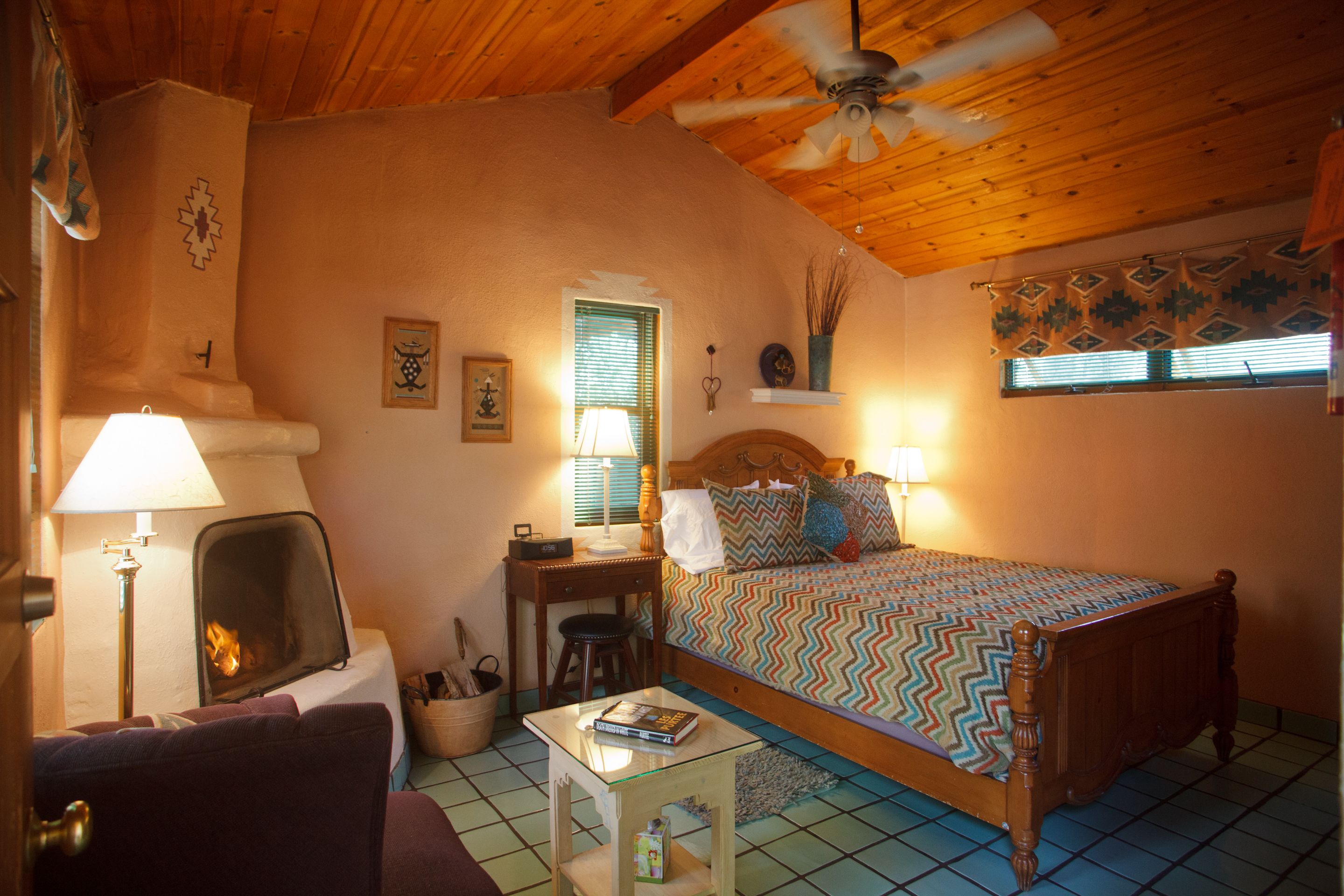 Une pièce chaleureuse avec une cheminée, un lit recouvert d’une couvre-lit à motifs, un bureau avec une lampe et un ventilateur de plafond.