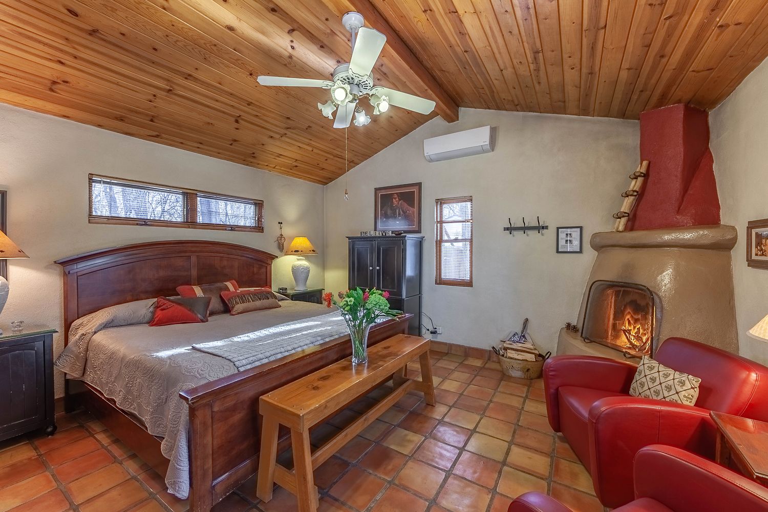 A cozy room with a wooden ceiling, a bed with a headboard, a wooden bench, a red leather chair, a fireplace, and a television.