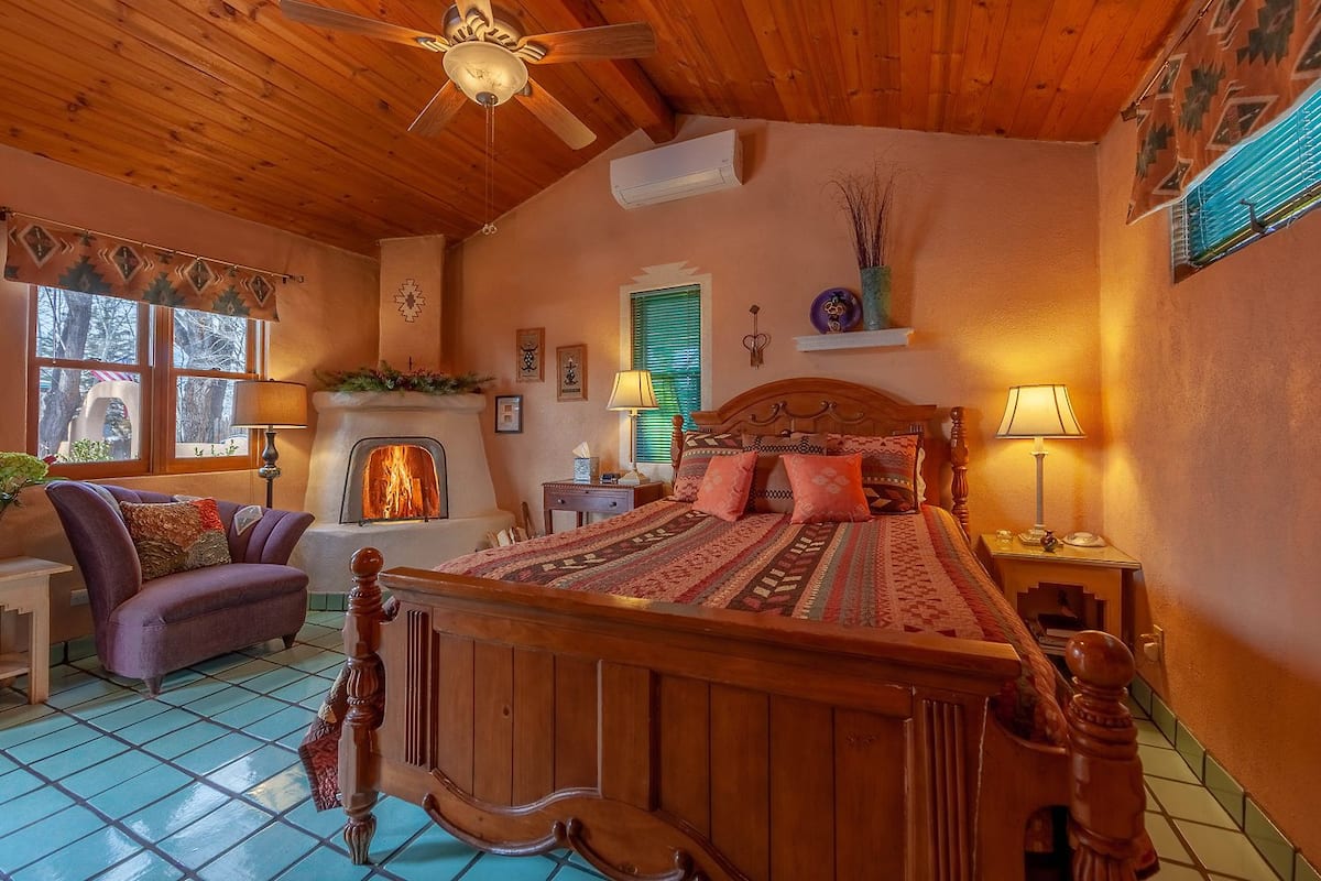 A cozy room with a wooden ceiling, a fireplace, a bed with patterned bedding, a purple armchair, and a small table with a lamp.