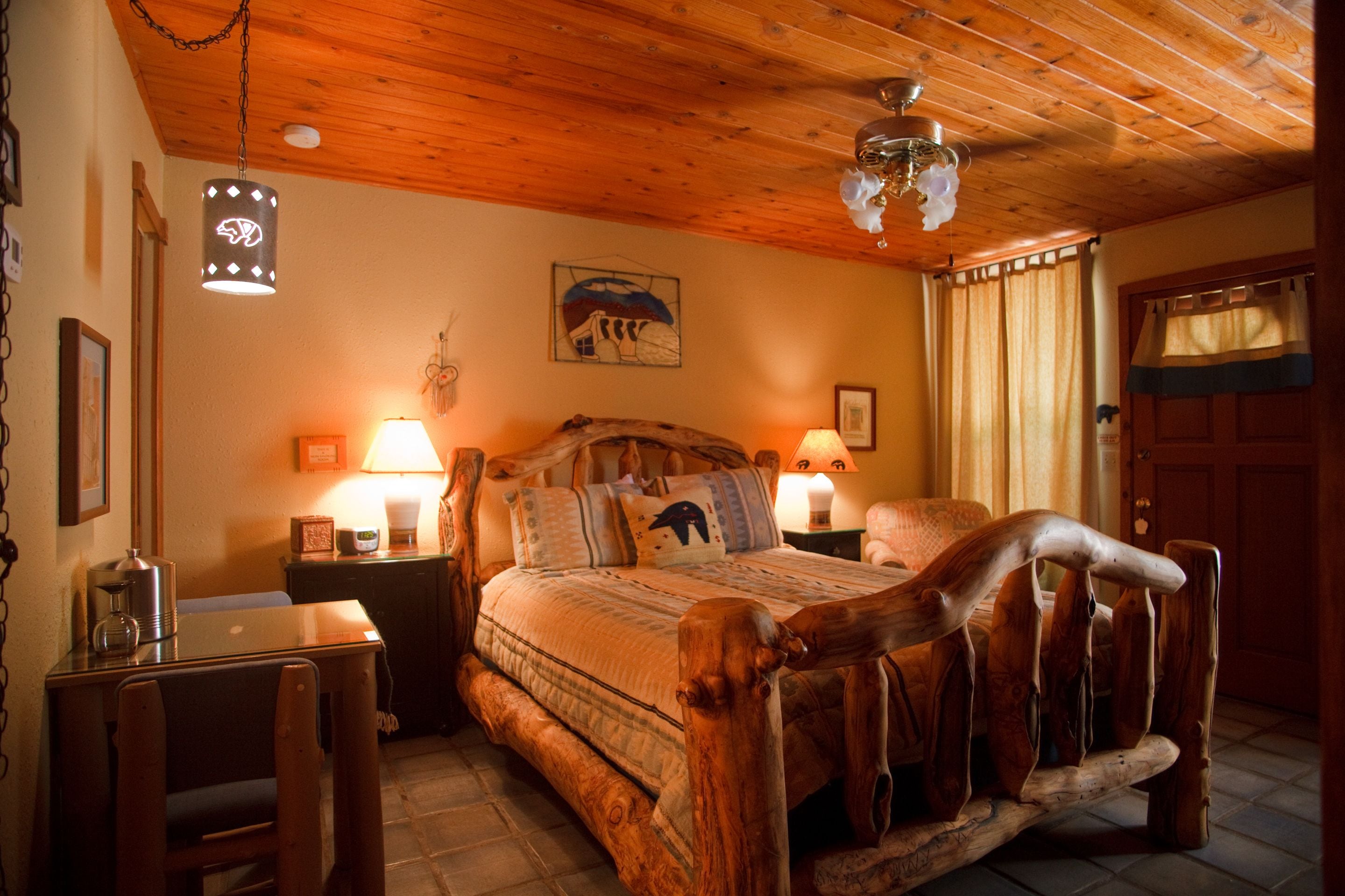 A rustic wooden bed with a headboard, a wooden table with a chair, and a hanging light fixture.