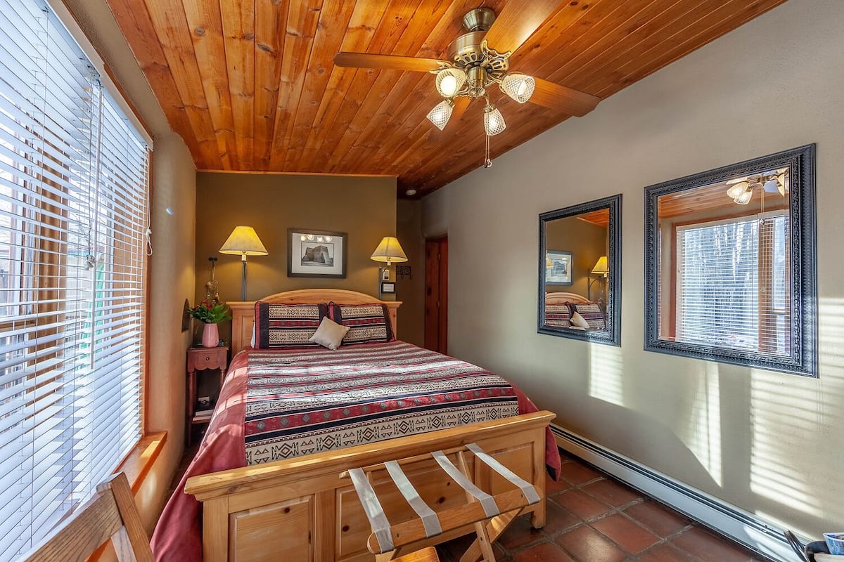 A bedroom with a wooden ceiling, a bed with patterned bedding, two bedside lamps, a ceiling fan, and a large mirror on the wall.