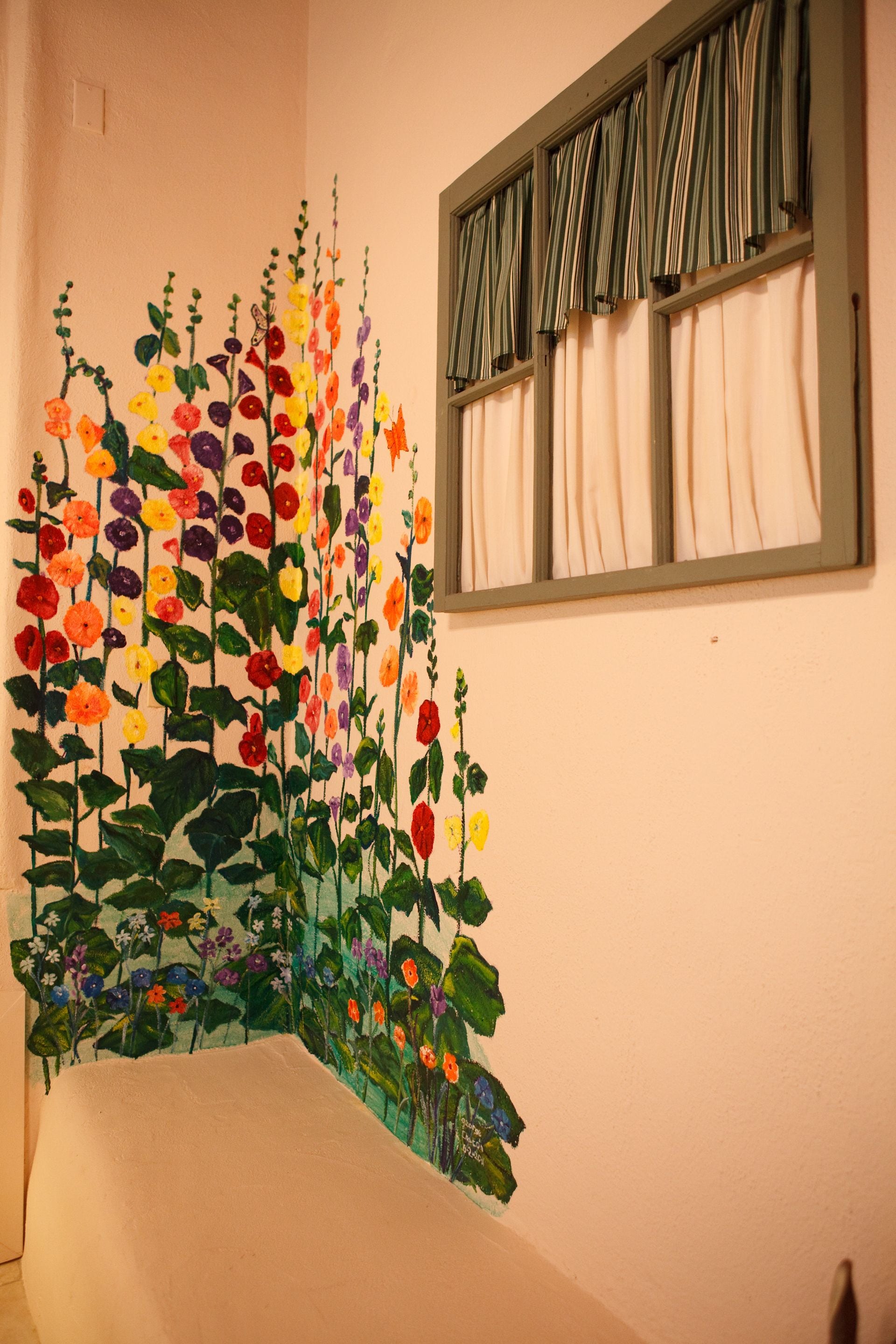 Una habitación con un cuadro floral grande en la pared, una ventana con cortinas y una cama con cabecera blanca.