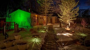 Een goed verlicht buitengebied met een houten terras, plantenbakken en een pad dat naar een gebouw leidt.