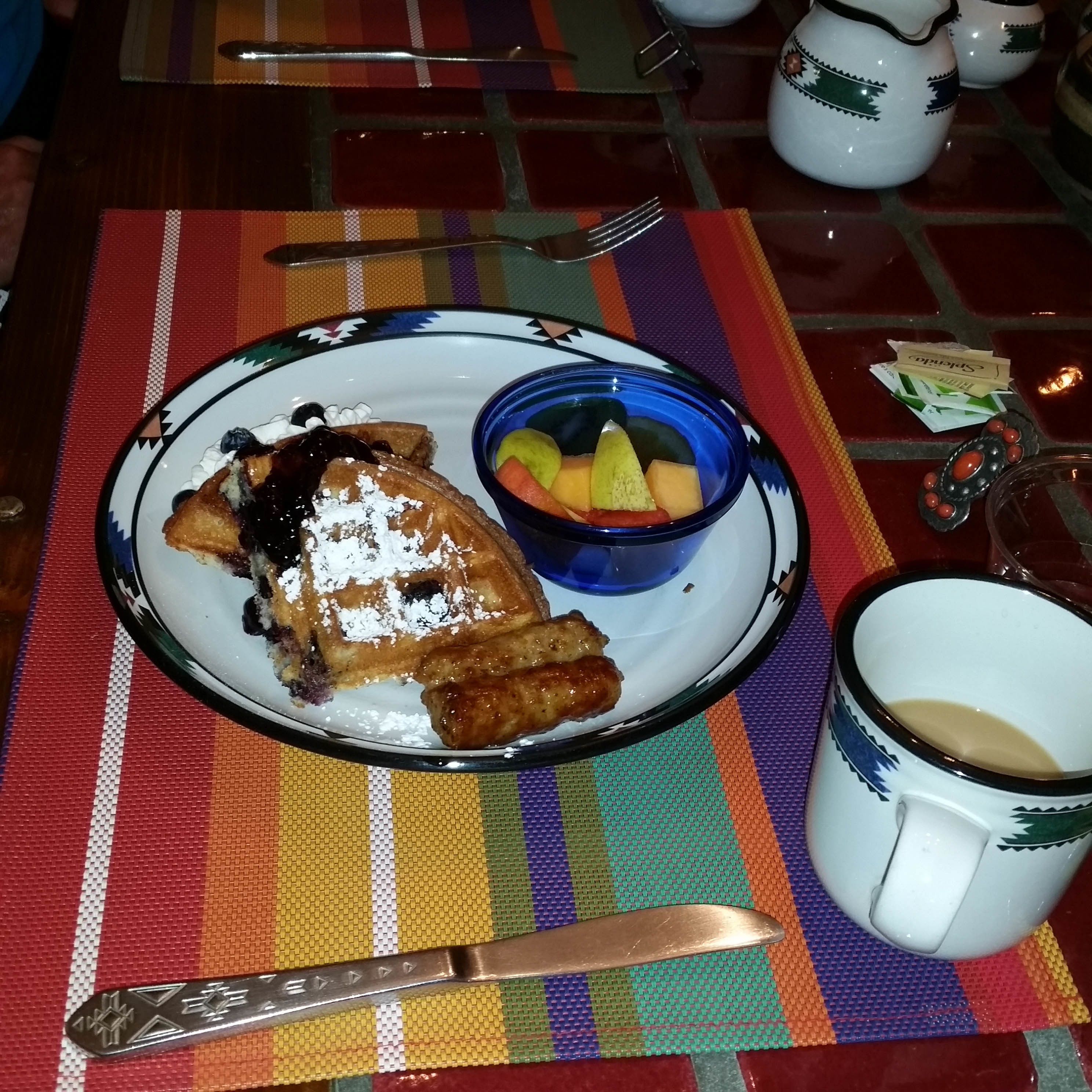 A breakfast plate with waffles, a fruit bowl, and a coffee mug on a colorful tablecloth.