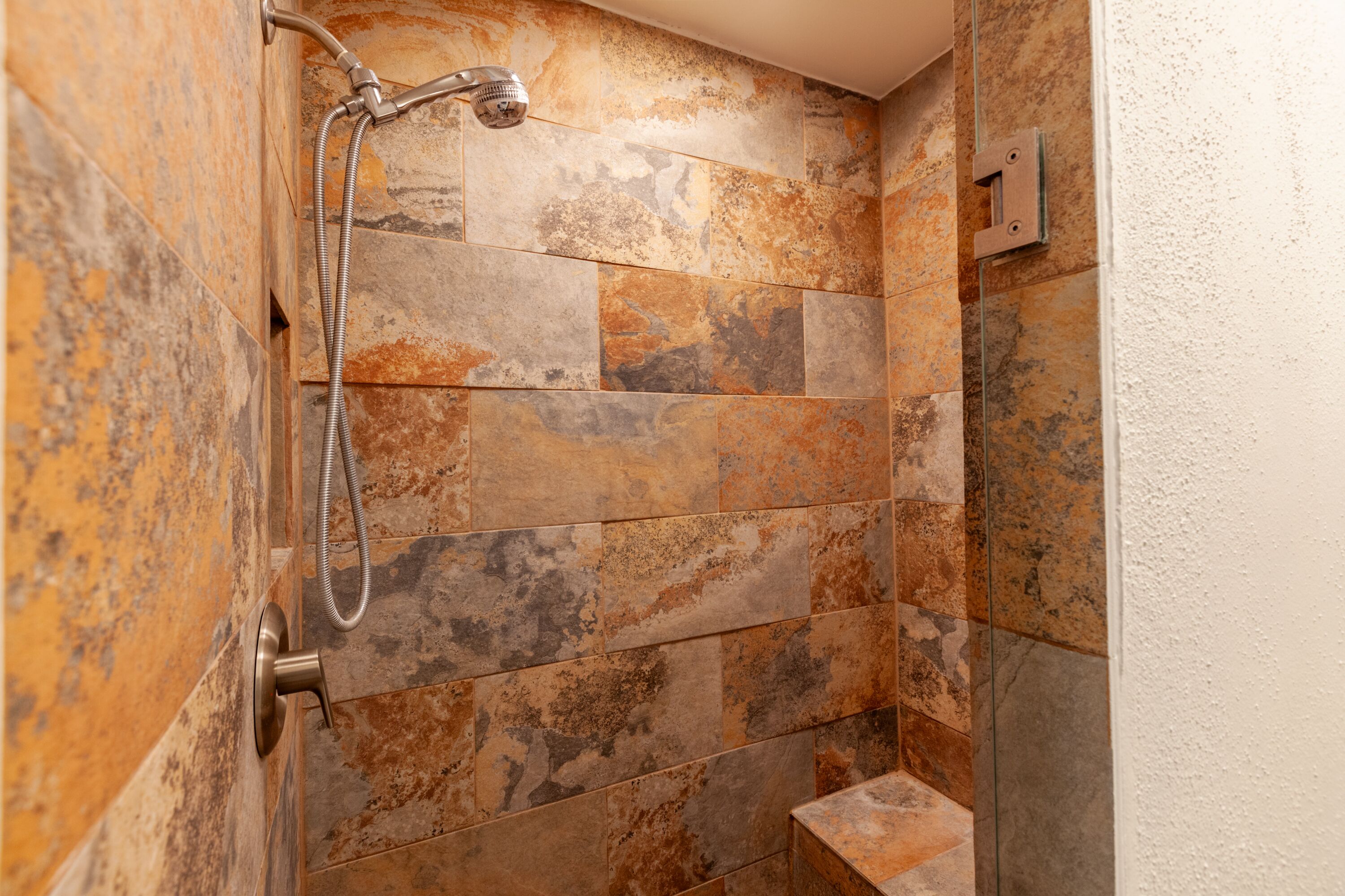 A shower with a handheld showerhead and a wall-mounted faucet, tiled with various shades of stone.