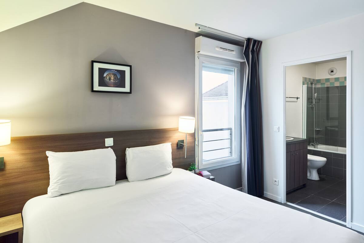 A hotel room with a large bed, a bedside table, a nightstand, a framed picture on the wall, a window with curtains, and a bathroom with a shower and toilet.
