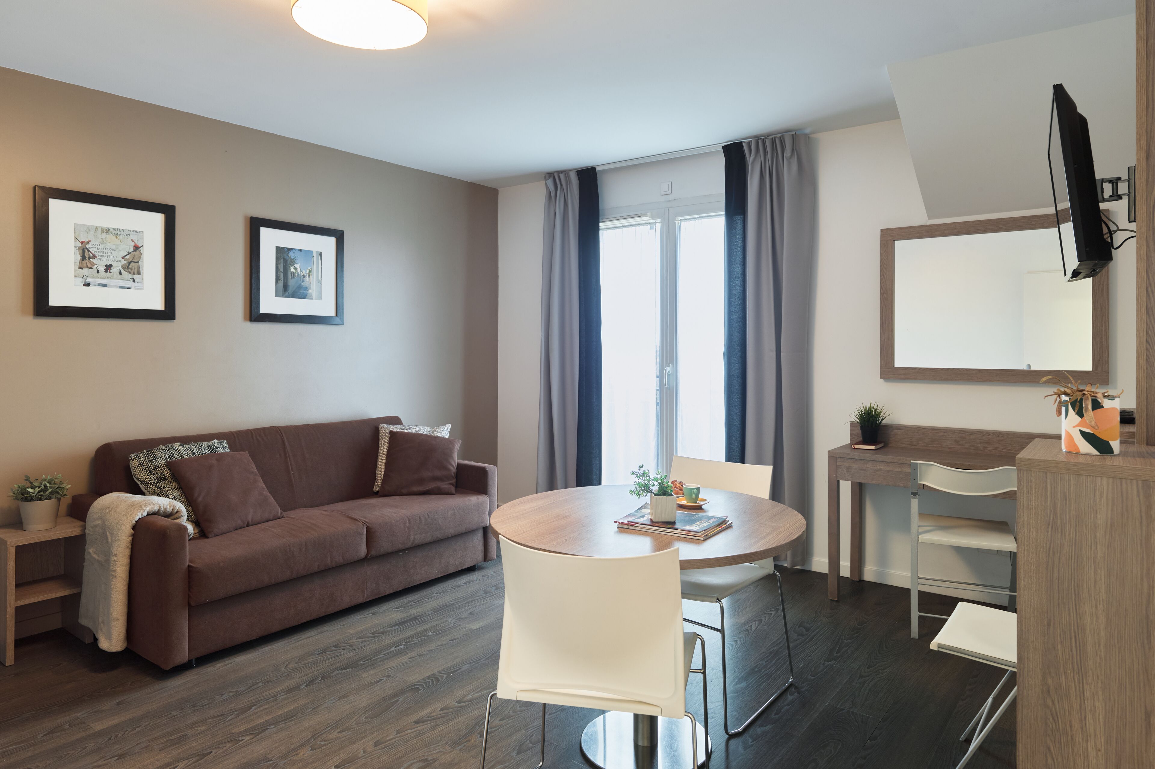 A modern living room with a brown sofa, a round dining table with chairs, a small side table, a TV mounted on the wall, and a window with curtains.
