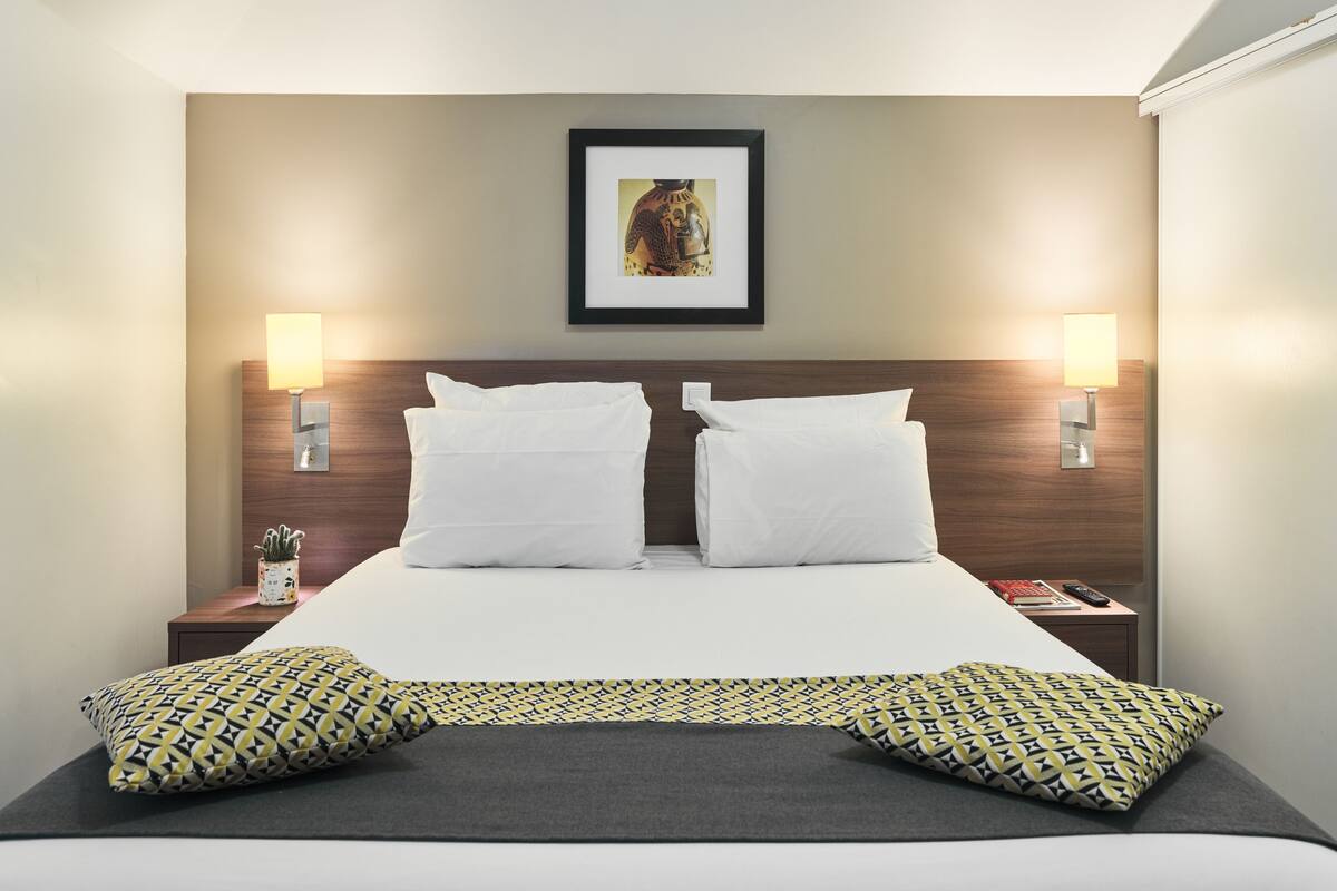 A hotel room with a large bed, two bedside lamps, a framed picture on the wall, and a small table with a plant.