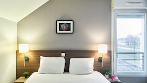 A hotel room with a neatly made bed, two bedside lamps, a framed picture on the wall, and a window with a view of the outside. - Zenitude Hôtel - Résidences Roissy Village (Roissy-en-France)