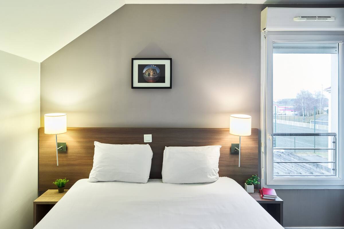 A hotel room with a neatly made bed, two bedside lamps, a framed picture on the wall, and a window with a view of the outside.