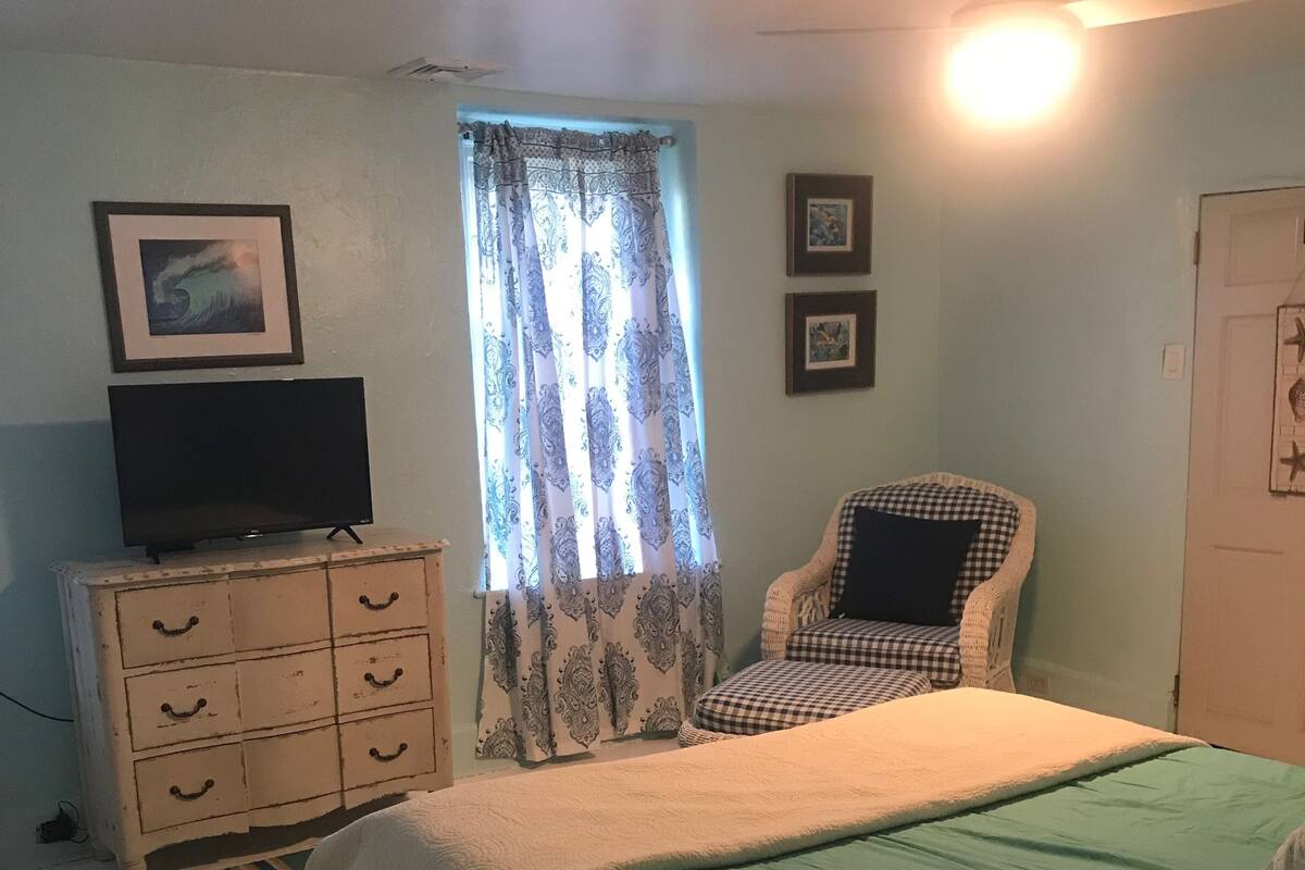 Une chambre à coucher avec un lit, une chaise, une commode, un téléviseur et une fenêtre avec des rideaux.