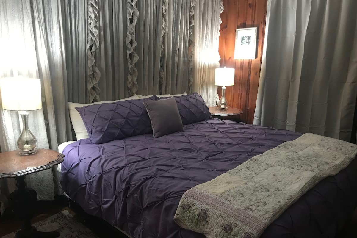 Un lit bien fait, recouvert d’une courtepointe violette, d’un plaid blanc et agrémenté d’une petite table de chevet avec une lampe.