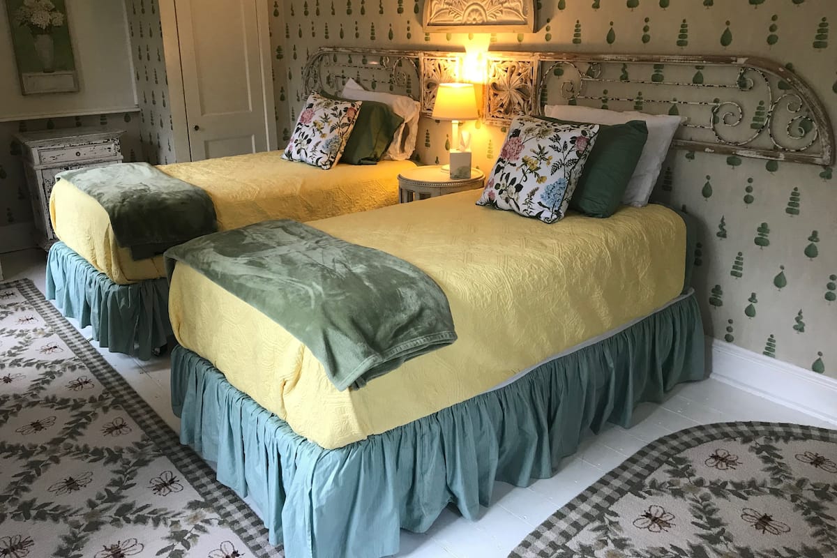 Une chambre avec deux lits, chacun recouvert d’une couvre-lit verte et jaune, assortie à des coussins du même style. Une table de chevet avec une lampe se trouve entre les deux lits.