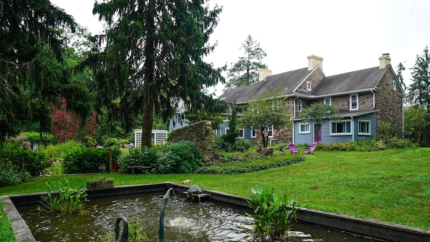 A pond with a statue, surrounded by a well-maintained garden and a stone house with a gabled roof. - Wild Wisteria (Chadds Ford)