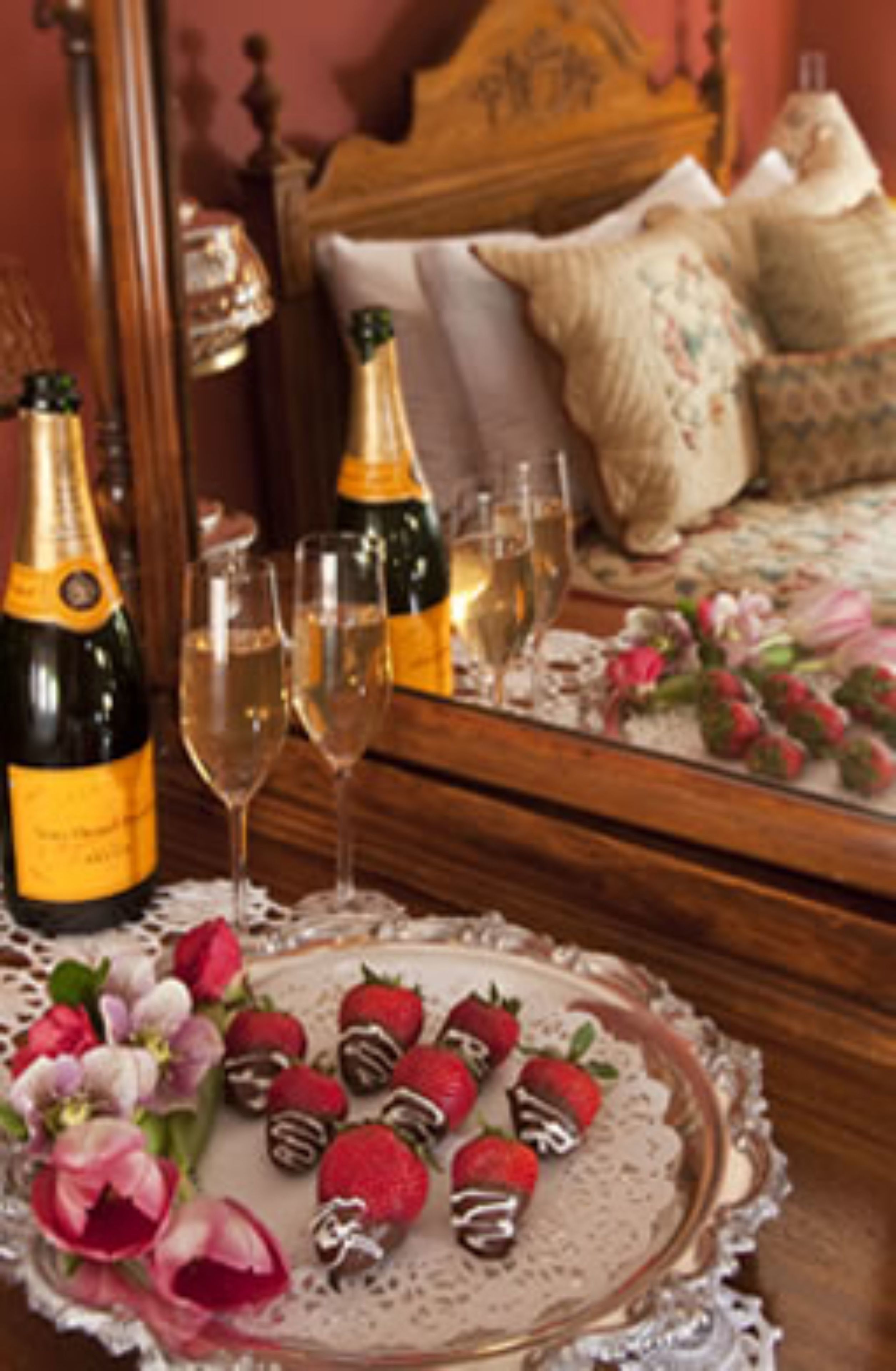A bedroom with a wooden bed, a bedside table with champagne bottles and glasses, and a tray with strawberries and flowers.