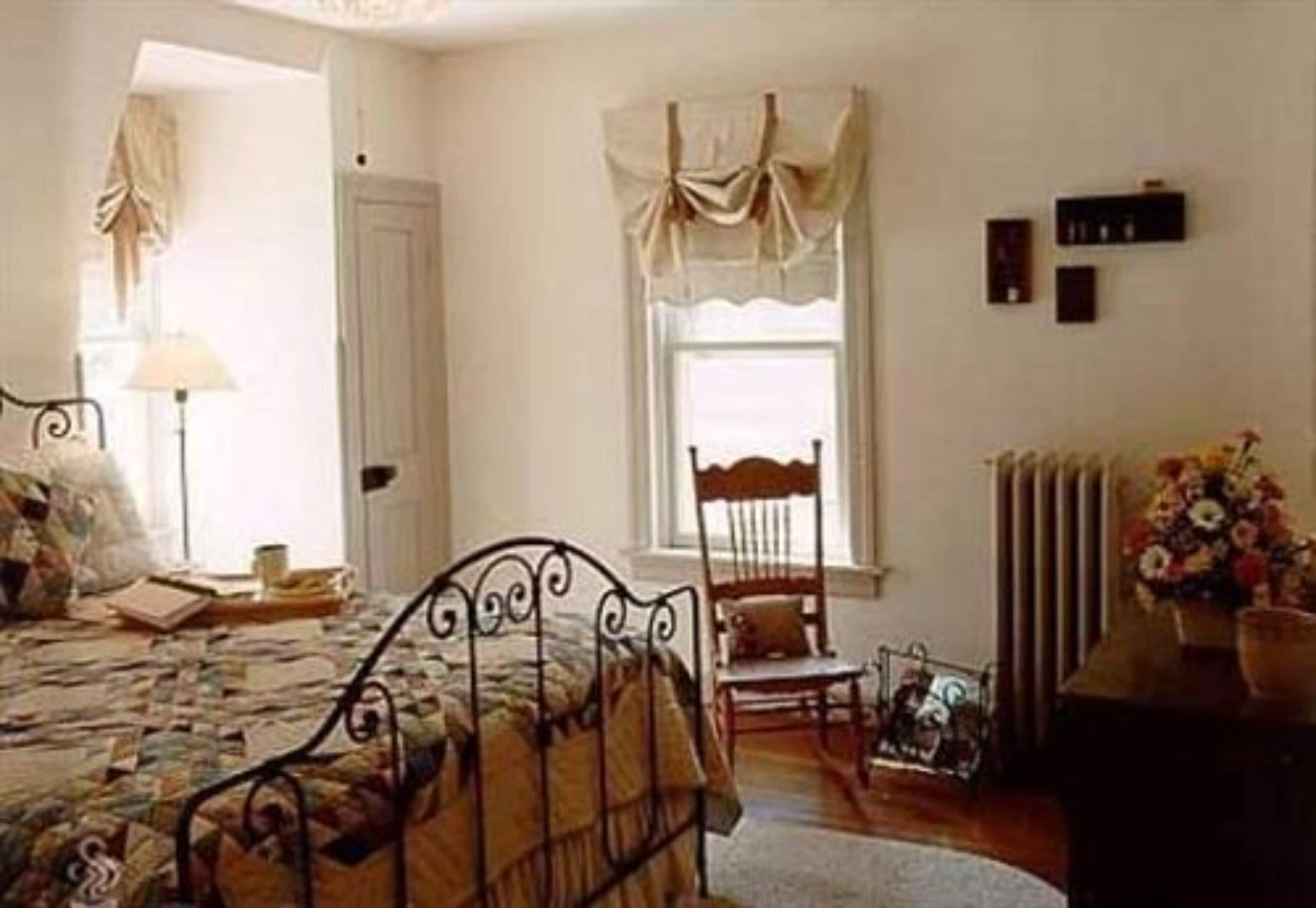 A bedroom with a metal bed frame, a wooden chair, a bedside table with a lamp, a window with curtains, and a radiator.