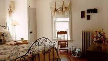 A bedroom with a metal bed frame, a wooden chair, a bedside table with a lamp, a window with curtains, and a radiator.