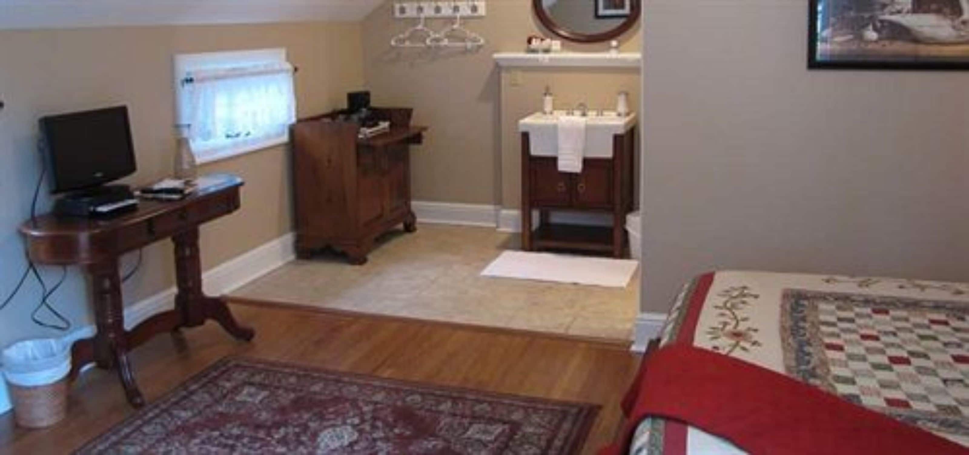 Quarto com cama, penteadeira, escrivaninha com computador, espelho, janela e banheiro com pia e espelho.